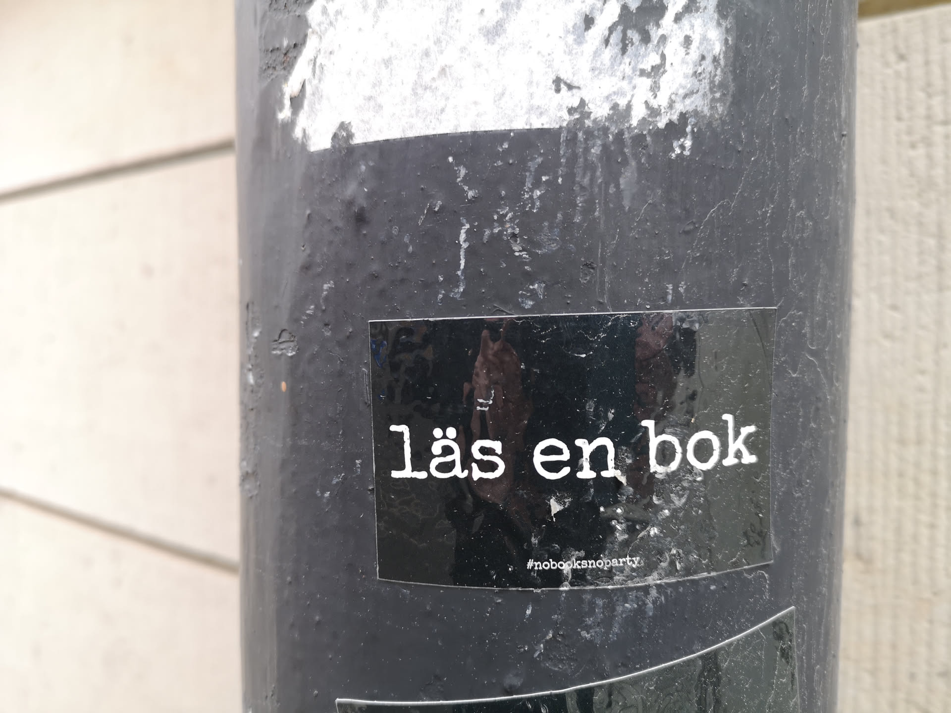 The sticker is rectangular with a glossy black background. It features white, sans-serif text in two lines. The main text, 'läs en bok', is larger and centered. Below it, in smaller font, is the hashtag '#nobooksno party'. The sticker shows some reflections from the surrounding environment, indicating its glossy surface and physical presence on a textured, dark grey pole.