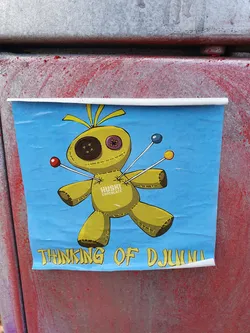 A voodoo doll sticker with a light blue background. The doll is yellow and has buttons for eyes. Pins with colored balls on top are stuck in it.  The text "HUSKI CHOCOLATE" is written on the doll. The words "THINKING OF DJLuigi" are written at the bottom.