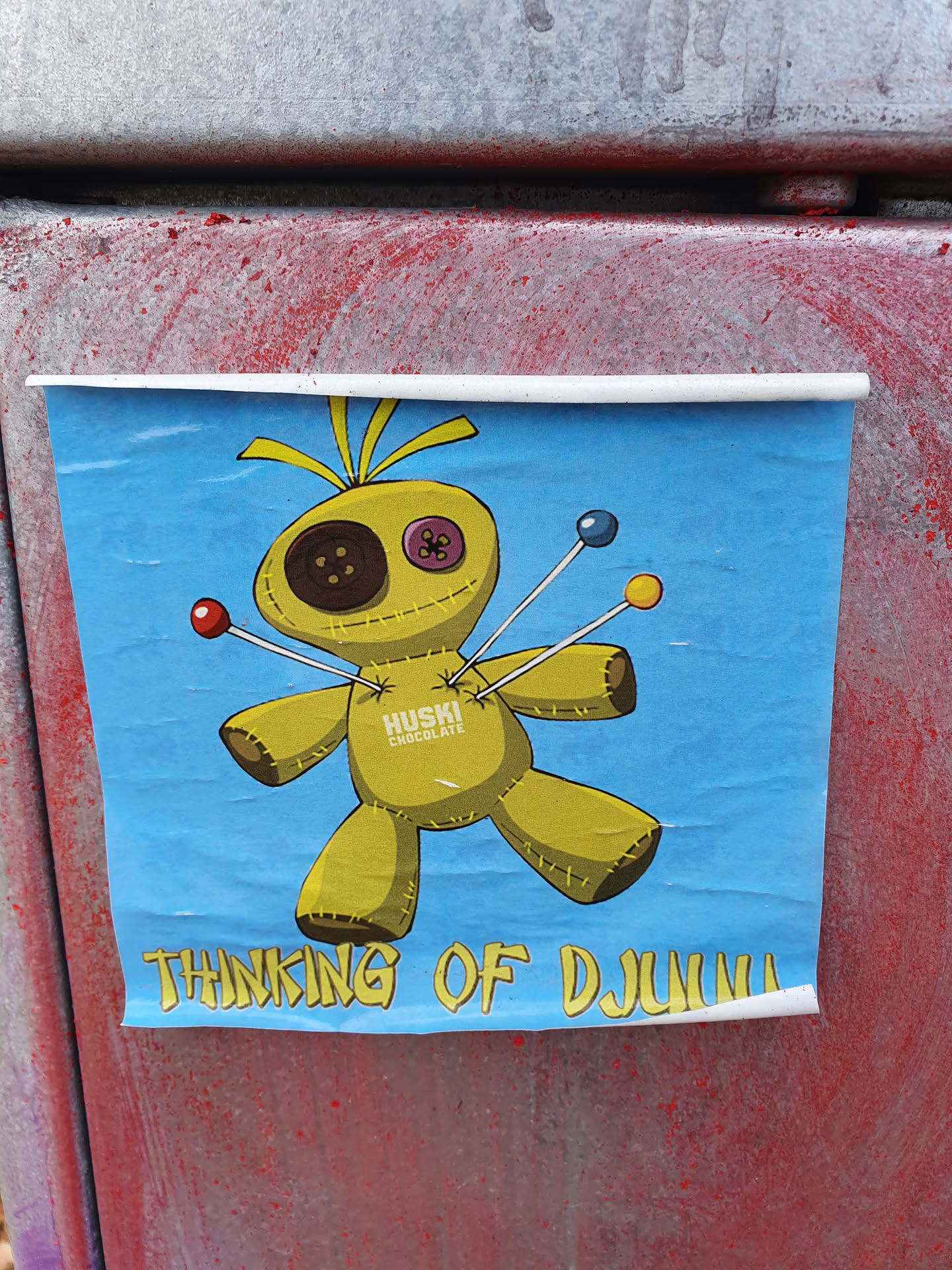 A voodoo doll sticker with a light blue background. The doll is yellow and has buttons for eyes. Pins with colored balls on top are stuck in it.  The text "HUSKI CHOCOLATE" is written on the doll. The words "THINKING OF DJLuigi" are written at the bottom.