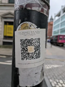 The sticker is a rectangular, white label affixed to a dark, weathered pole. It features black sans-serif text at the top, a large black and white QR code in the center, and an email address at the bottom. The QR code has a small, light brown and orange logo embedded in its center, which reads 'INTUITION COACH' below a stylized, circular, dotted pattern. The sticker shows some minor creasing and dirt, indicating it's been exposed to the elements.