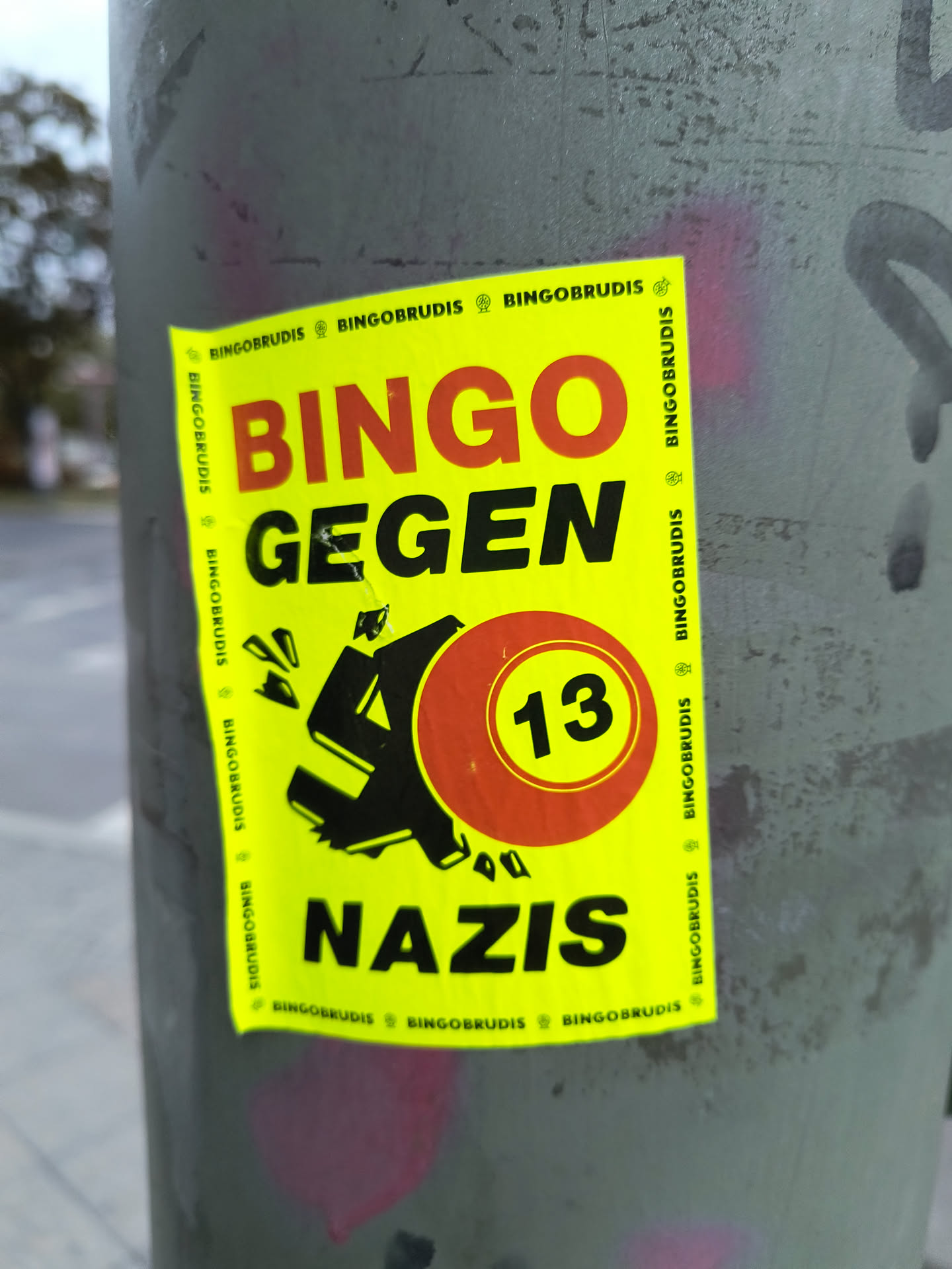 The sticker is bright yellow with bold red and black lettering. The main text reads 'BINGO GEGEN NAZIS'. In the center, there is a stylized black symbol resembling a broken swastika, with fragments flying off. To the right of this symbol is a red circle with the number '13' in yellow. The edges of the sticker have a repeating pattern of the word 'BINGOBRUDIS' with a small symbol between each instance.