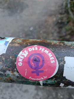 Pink and purple feminist sticker with a raised fist inside a Venus symbol. The text on the sticker says "Grève des femmes" which translates to "Women's Strike" in English and the website address Fræetraik.lu. The sticker is affixed to a rusty metal surface.