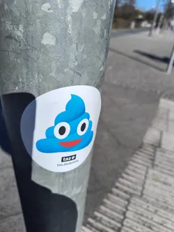 A round sticker on a pole features a bright blue poop emoji with a cheerful red smile and large white eyes with black pupils. Below the emoji is a small square containing the word "taz" in white text on a black background. Underneath this is the url taz.de/antifa.