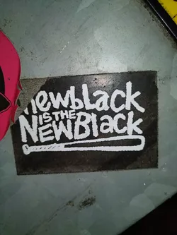 A black and white sticker with the text "New Black is the New Black" and an image of a baseball bat. The sticker is affixed to a dirty surface.
