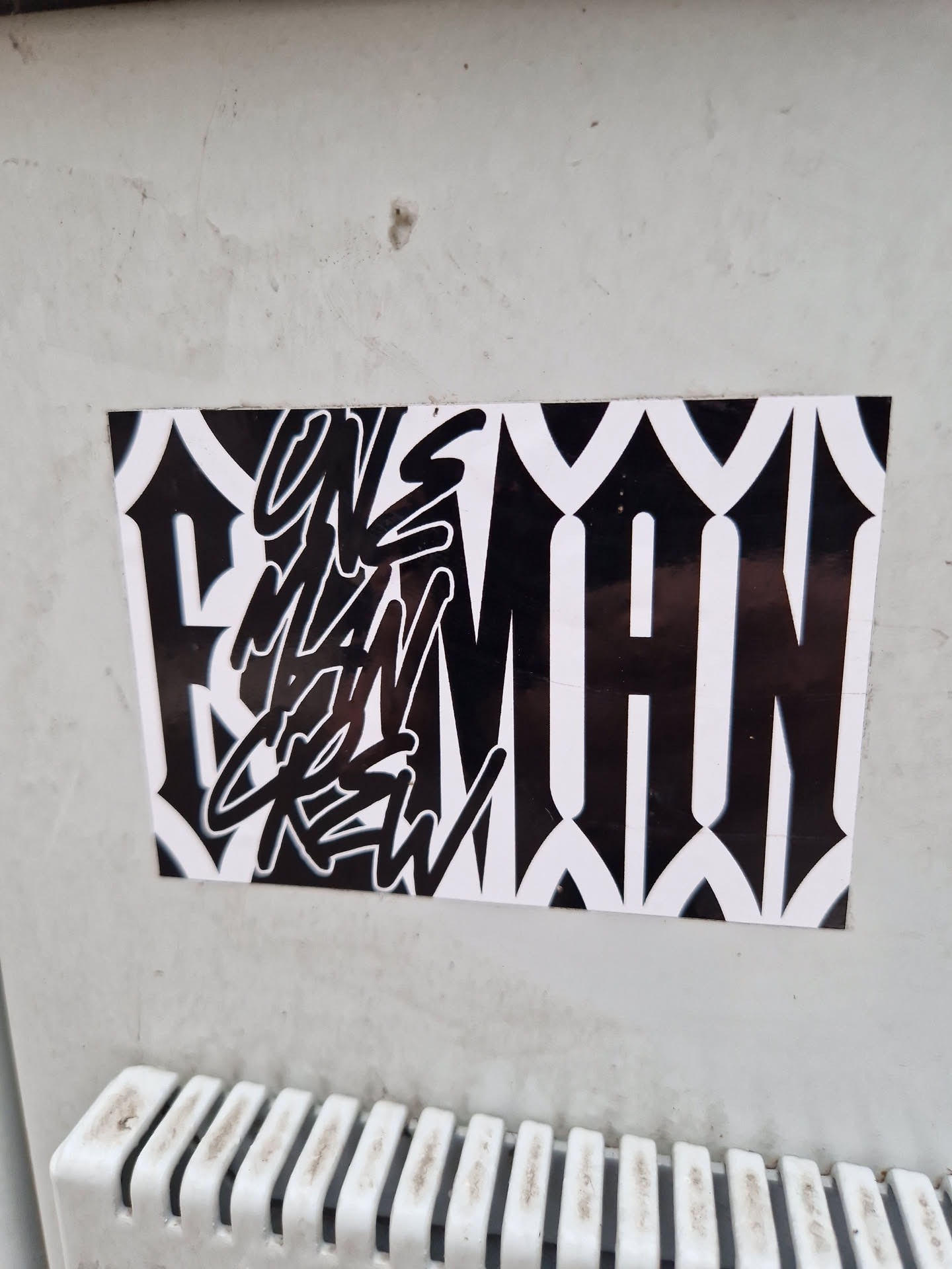 Black and white sticker with the text "Uns Man Crew" in a stylized graffiti font. The background features a geometric pattern.