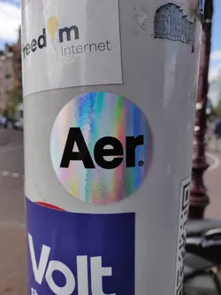 A round holographic sticker with the text "Aer" in black uppercase letters on a rainbow iridescent background. The sticker is affixed to a pole with other stickers.