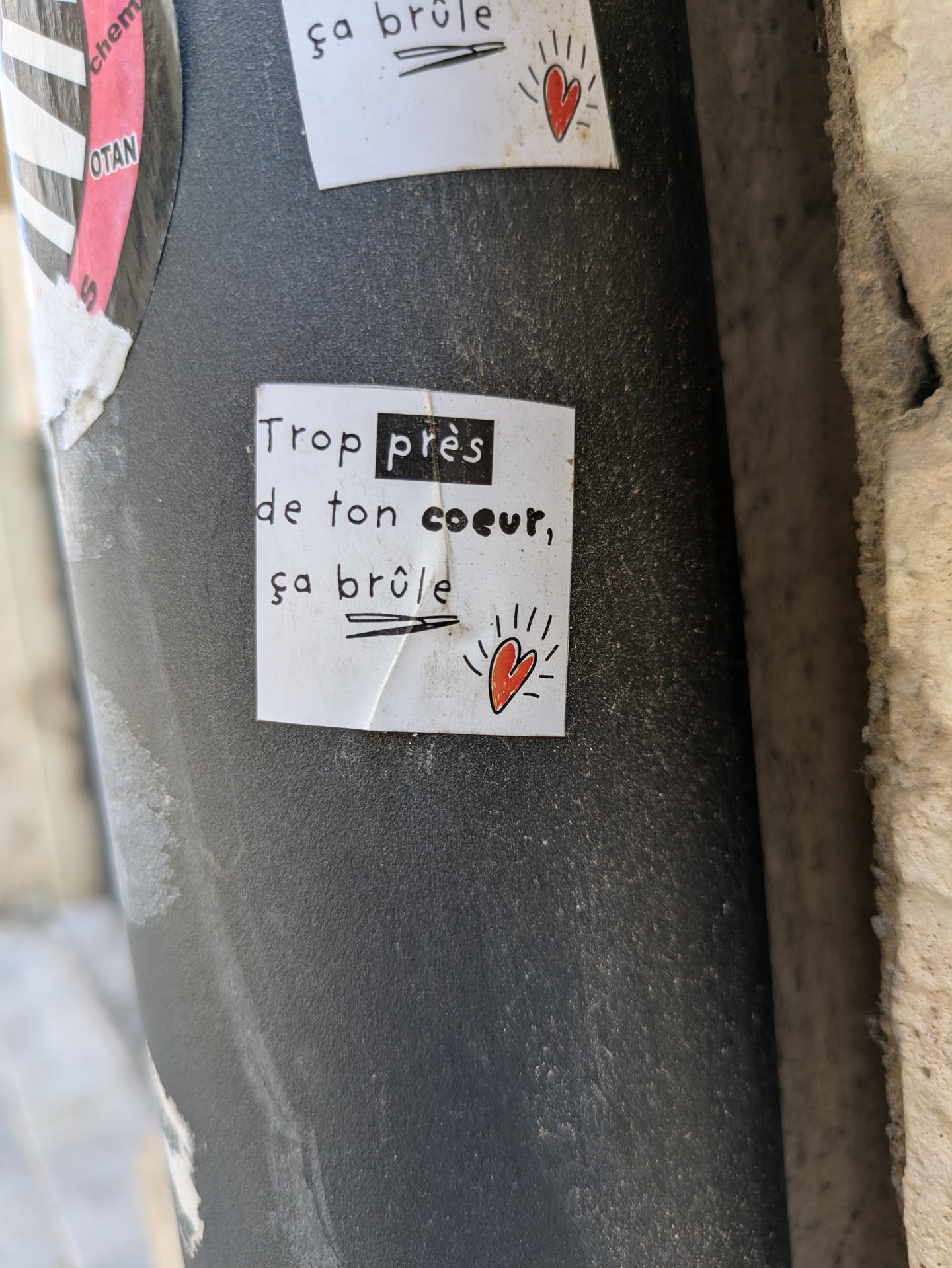 A small, square sticker on a pole reads "Trop près de ton coeur, ça brûle" (Too close to your heart, it burns). A small red heart is drawn near the bottom right corner of the sticker.