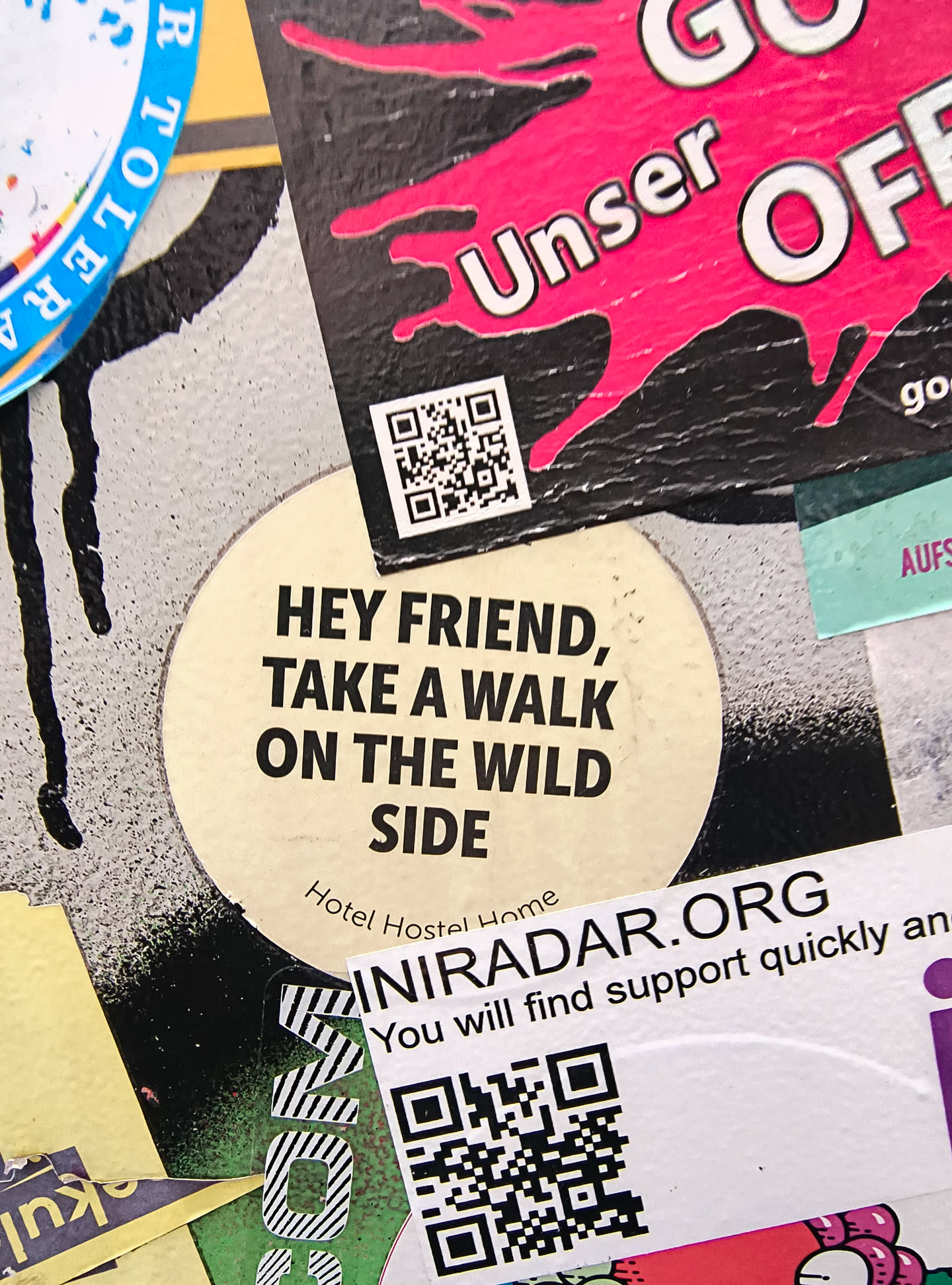 A circular, cream-colored sticker with bold, black sans-serif text. The text reads 'HEY FRIEND, TAKE A WALK ON THE WILD SIDE'. Below this, in smaller text, it says 'Hotel Hostel Home'. The sticker has a slightly aged appearance with some subtle speckling and a faint shadow effect around the edges.