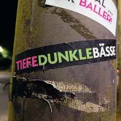A black sticker with the text "Tiefe dunkle Bässe" in pink and light green. The sticker is attached to a pole.