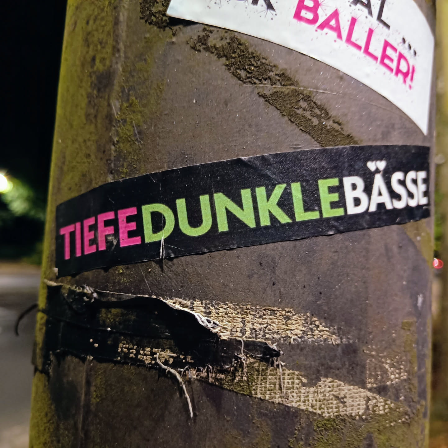 A black sticker with the text "Tiefe dunkle Bässe" in pink and light green. The sticker is attached to a pole.