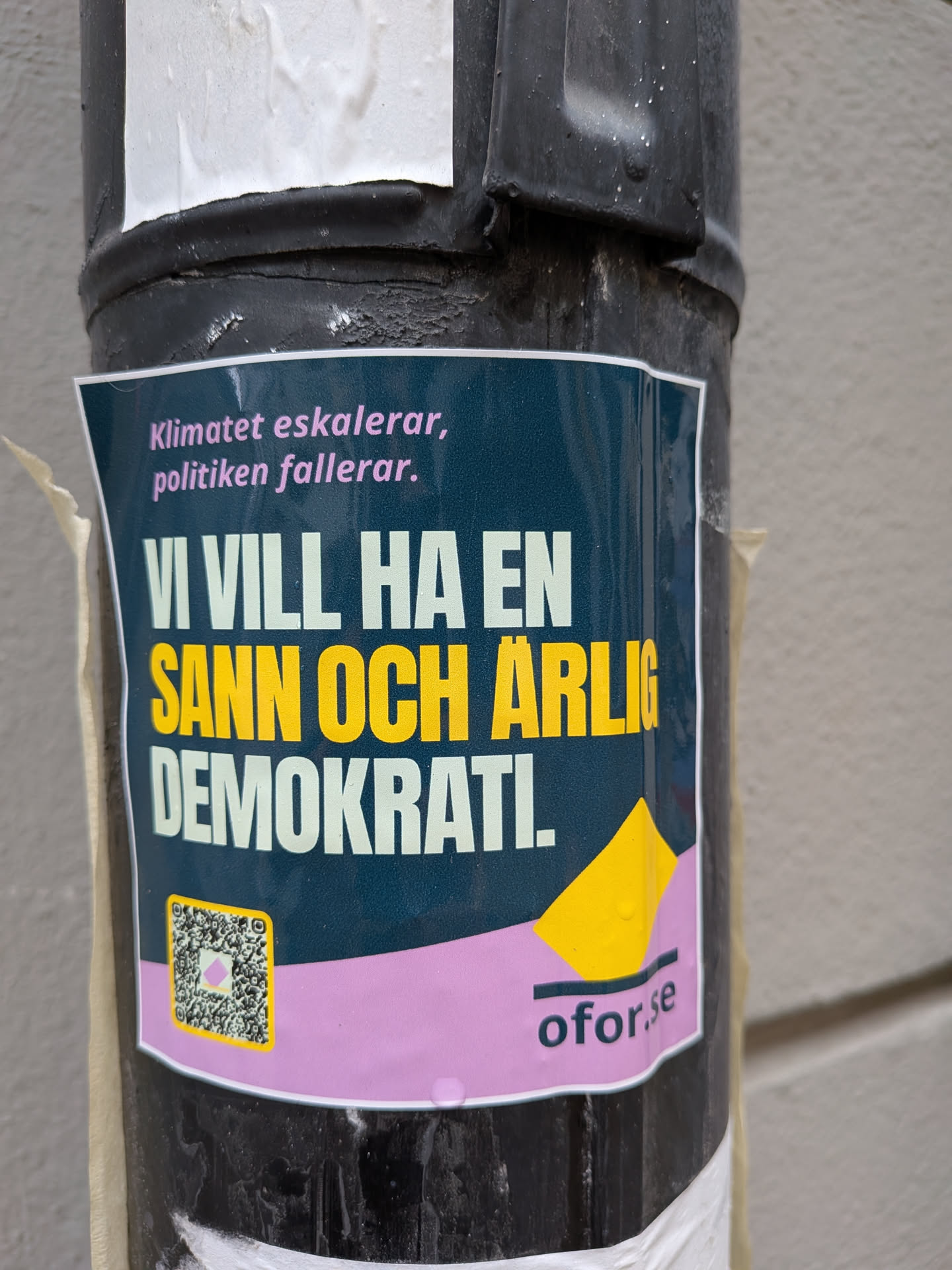 A rectangular sticker affixed to a black pole, featuring a dark teal/blue background. The top section displays the text 'Klimatet eskalerar, politiken fallerar.' in pink. Below this, the phrase 'VI VILL HA EN' is prominently displayed in white, followed by 'SANN OCH ÄRLIG DEMOKRATI.' in bright yellow, all in a bold, sans-serif font. The bottom portion of the sticker is a light pink/purple band. On the left of this band, there is a QR code with a yellow border and a pink diamond in its center. To the right, a yellow abstract, folded-paper-like shape is visible, next to the website 'oför.se' written in dark teal/blue. The sticker shows signs of outdoor exposure, with visible water droplets and slight wrinkling.