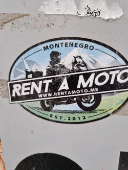 This sticker is for Rent a Moto, a motorcycle rental company based in Montenegro.  The sticker features a graphic of a motorcycle rider in front of a mountain landscape, along with the company's website and establishment year.