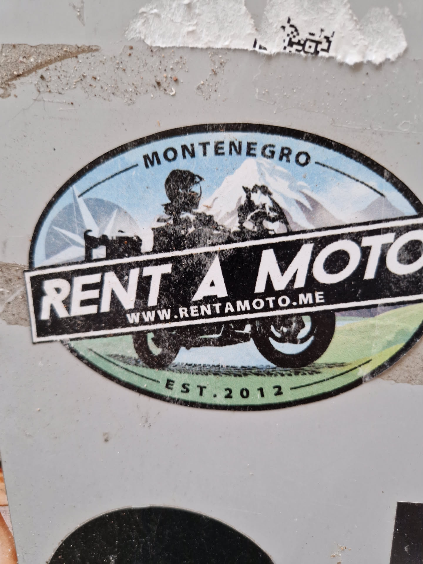 This sticker is for Rent a Moto, a motorcycle rental company based in Montenegro.  The sticker features a graphic of a motorcycle rider in front of a mountain landscape, along with the company's website and establishment year.