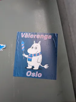 Street sticker A square paper sticker on a dark blue background featuring the classic Finnish character Moomintroll. Moomin is depicted smiling, wearing a blue and reddish-purple striped scarf, and holding a small firework rocket with matching colors. The text 'Vålerenga' is printed in outlined pinkish font along the top, and 'Oslo' is printed in the same style along the bottom.