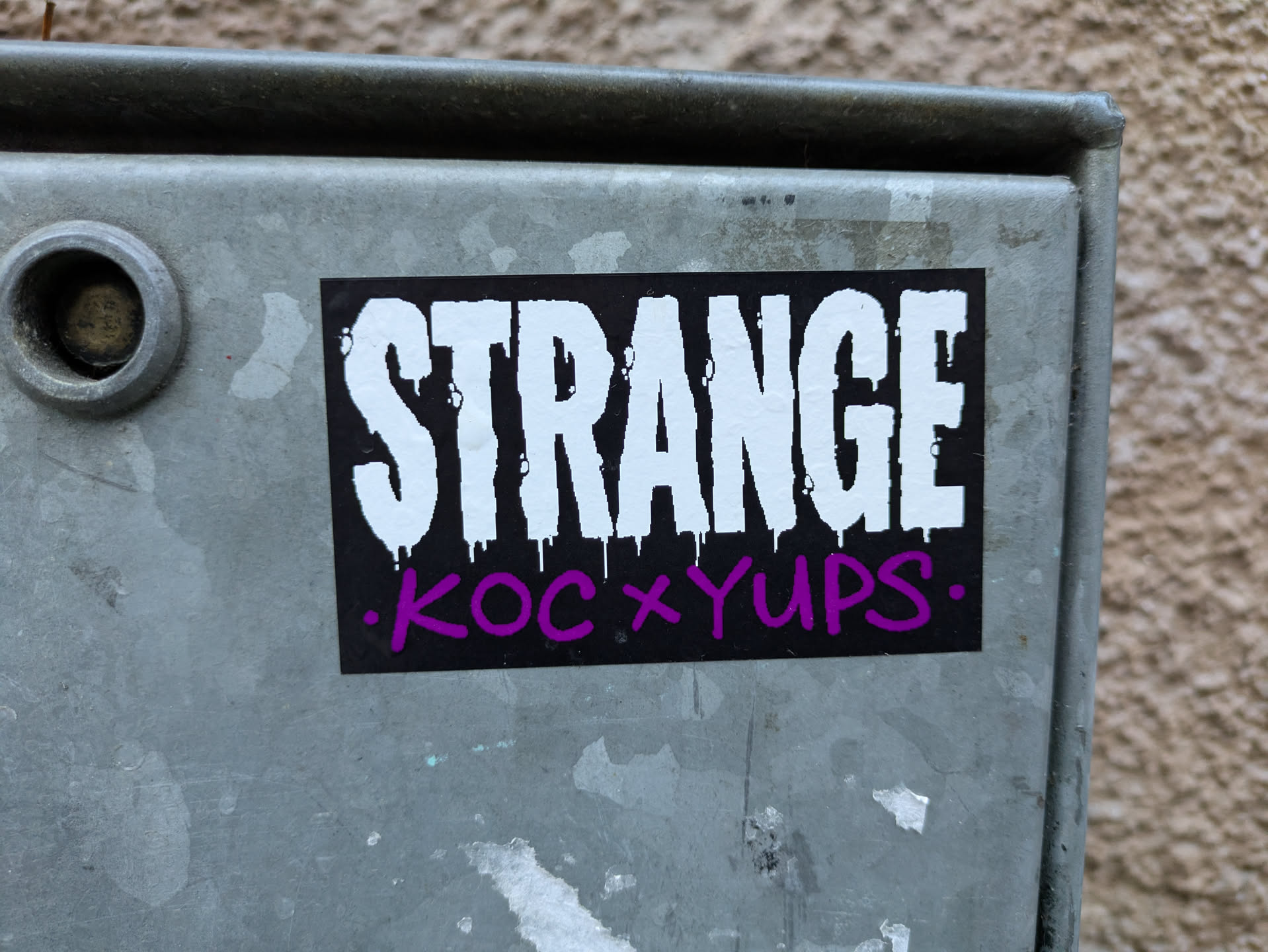 A black sticker with white dripping text that says "STRANGE" and purple text that says ".KOC x YUPS." The sticker is placed on a grey metal surface.