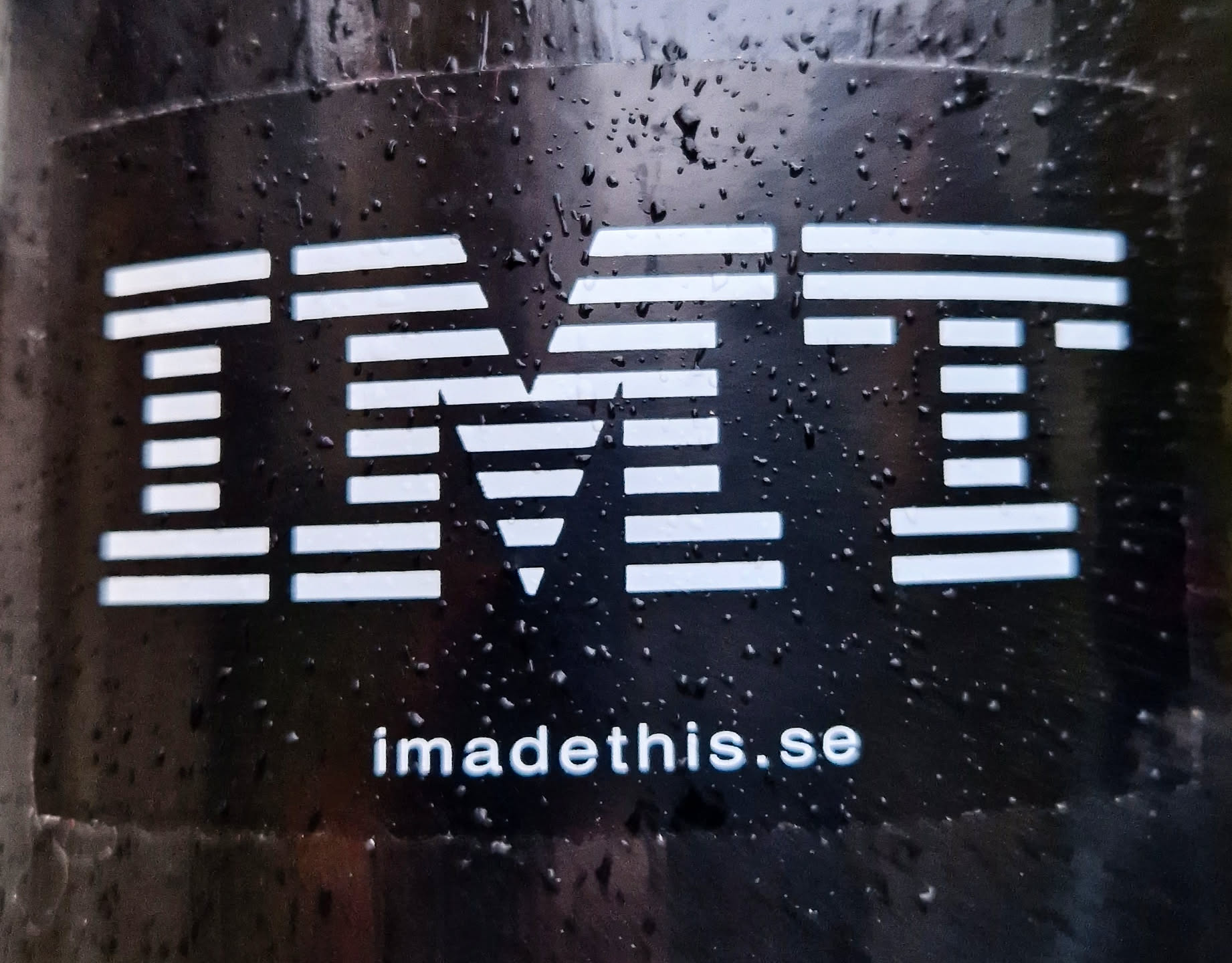 A black sticker with a white IMT logo. The letters are made up of horizontal lines. Below the logo, the website 'imadethis.se' is printed in white.