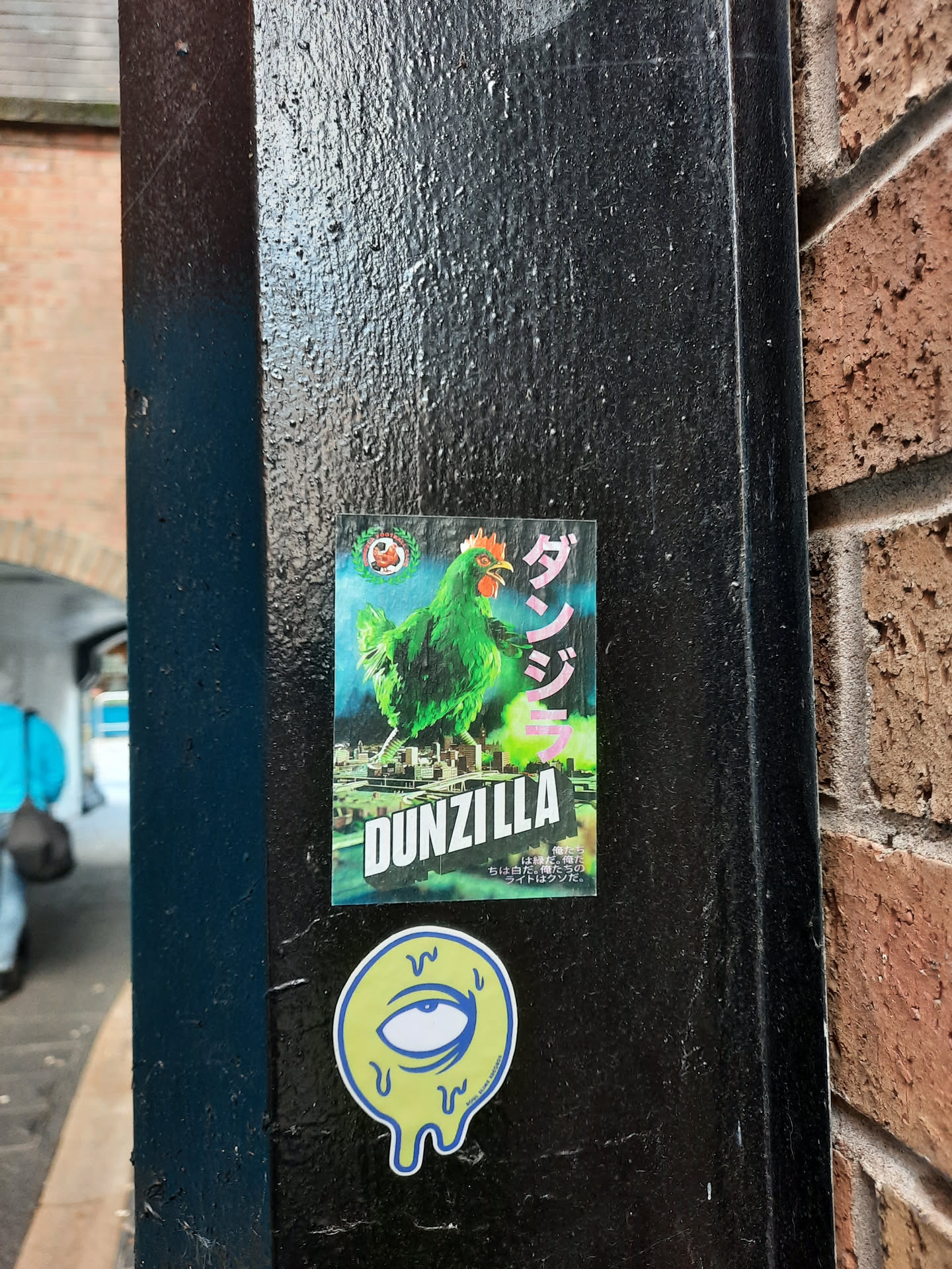 The central sticker features a vibrant green chicken, depicted in a kaiju-like pose, standing over a stylized cityscape. The chicken has a red comb and wattle, and its wings are spread. Behind it, the city appears to be under a dramatic, possibly stormy, sky with hints of pink and blue. To the right of the chicken, large Japanese characters are visible in pink. Below the chicken, the word 'DUNZILLA' is prominently displayed in bold, white, block letters with a green outline. Smaller Japanese text is present at the bottom of this sticker. Below this main sticker is a smaller, circular sticker with a yellow background and a blue outline. It features a stylized eye with a blue iris and pupil, surrounded by dripping lines, giving it a melting appearance. The eye sticker also has some faint blue text around its bottom edge.