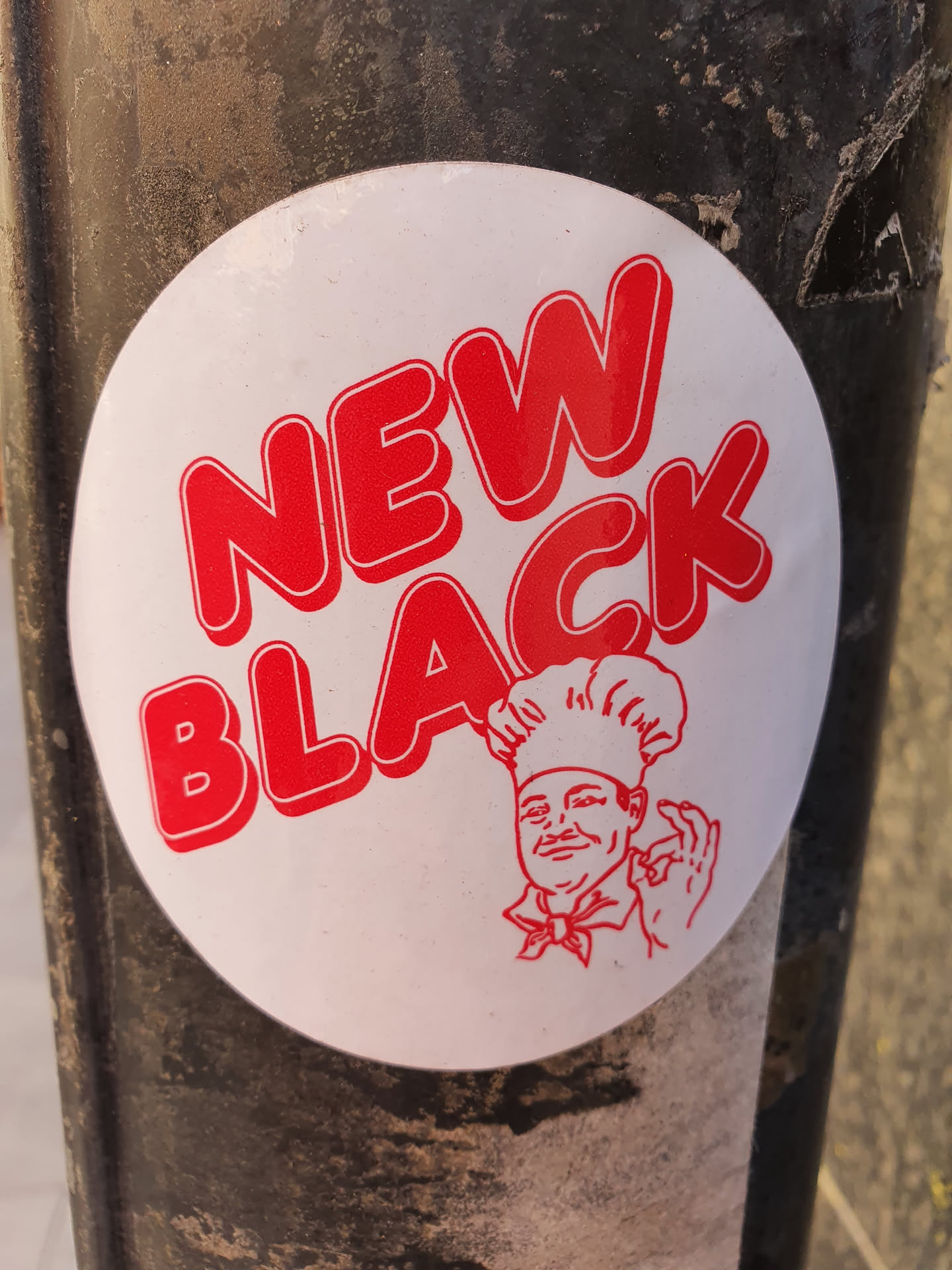 A round white sticker with red text and an illustration of a chef. The text says "NEW BLACK" in a bold, stylized font. The chef is depicted wearing a chef's hat and uniform, and is giving an okay sign.