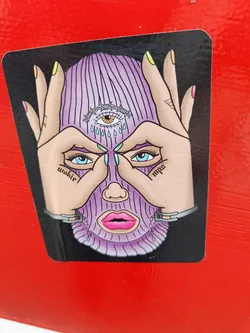 A sticker depicting a stylized face wearing a purple balaclava, with hands framing the face in a 'framing' gesture. The balaclava has an eye with a barbed wire crown on the forehead, and the words 'woke' and 'woke' are written on the sides. The style is colorful and bold, with a slightly edgy vibe.