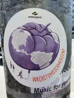 Street sticker A round sticker with an illustration of the earth as a purple balloon being inflated by a small person. The text "Wachstumsschmerzen?" is visible at the bottom, which translates to "growing pains?" in English.  The sticker also features the logo of Bundjugend at the top.