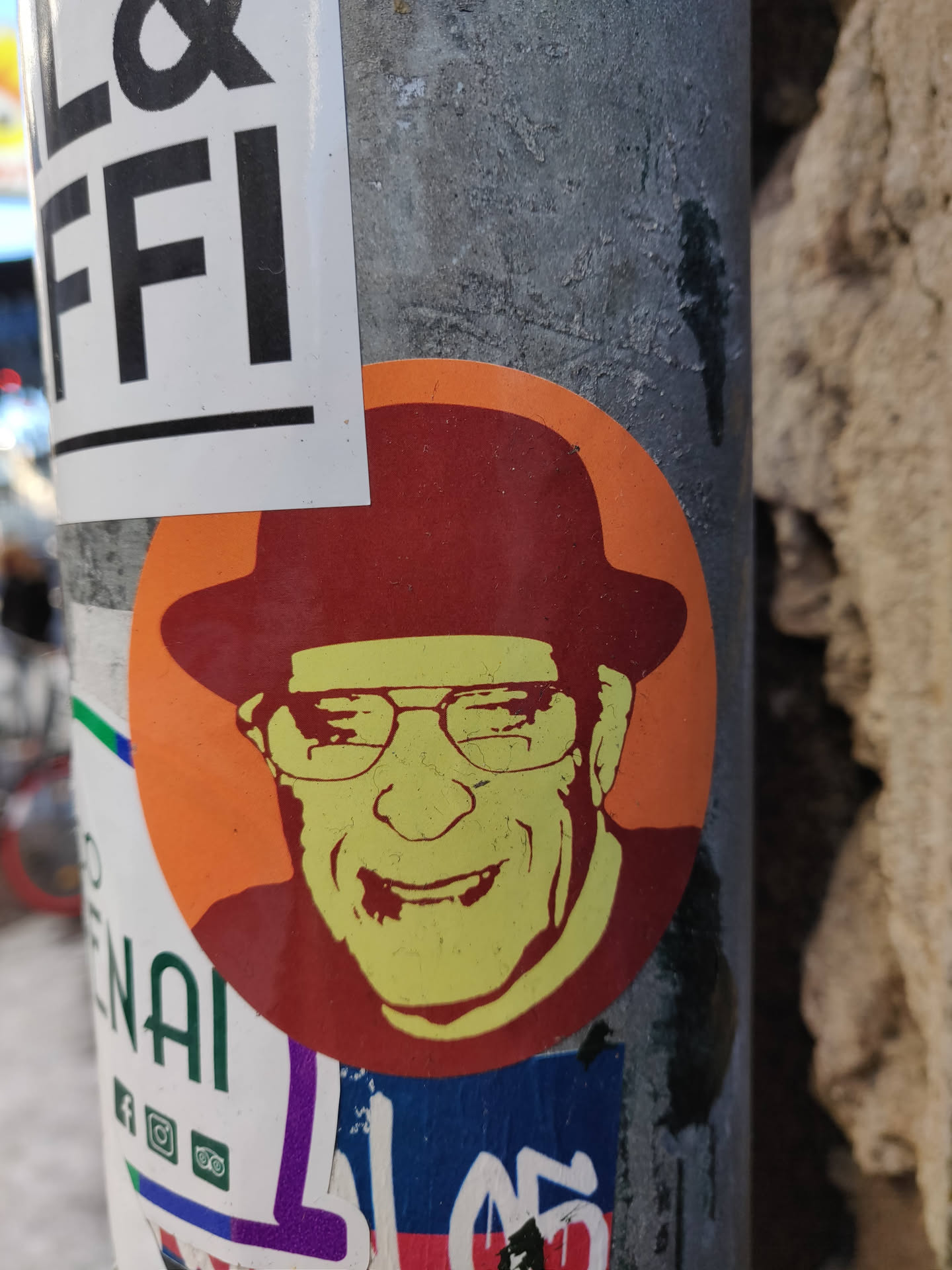 This round sticker showcases a vibrant, almost pop-art-like portrait of a smiling man. He's depicted wearing a fedora and glasses, giving him a classic, retro feel. The color palette is bold and striking, with the main image in shades of orange, yellow, and red against a contrasting background. The sticker's style is simple yet eye-catching, perfect for adding a touch of personality to any surface.