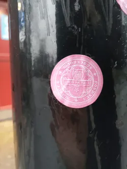 A pink, circular sticker is affixed to a dark-colored pole. The sticker features an illustration of an owl within an ornate design.  Surrounding the owl is text that appears to be reversed or in a mirror font, making it difficult to read completely. 