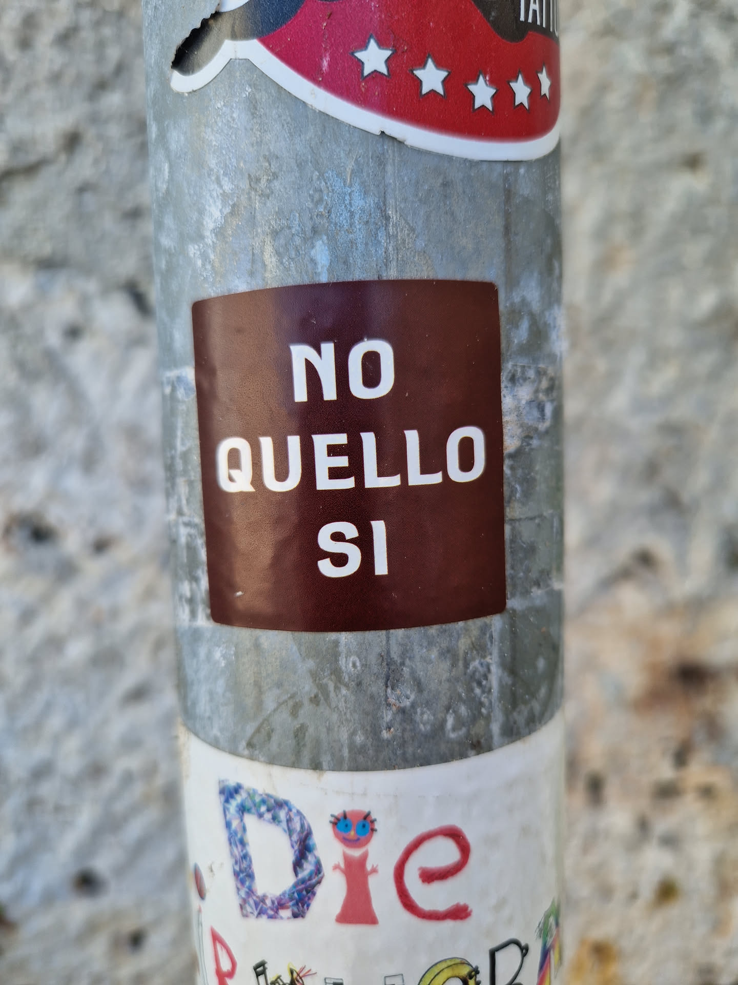A brown square sticker with the text "NO QUELLO SI" on it,  attached to a metal pole. The text is in Italian and translates to "No That Yes".