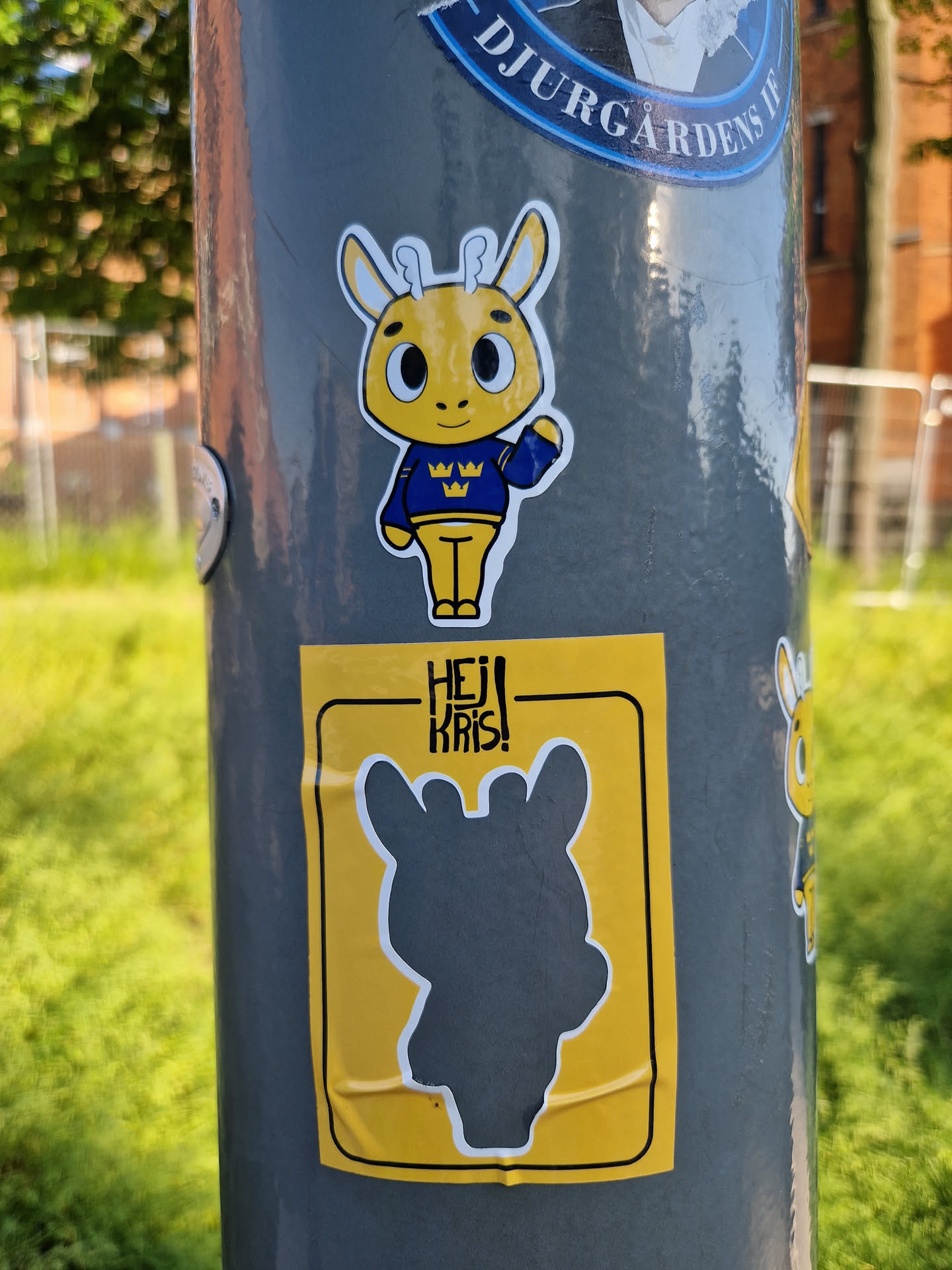A pair of stickers featuring a yellow deer mascot. The top sticker shows the mascot in a blue sweater with a logo on it.  The bottom sticker is a yellow square with a grey silhouette of the same deer and the text 'HEJ KRIS!'