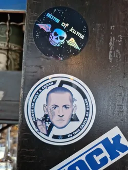 A round sticker featuring a portrait of Chester Bennington, lead singer of Linkin Park. The text surrounding the image reads, "Cause I'm only a crack in this castle of glass."  There is also a smaller sticker featuring a skull with wings and the text "Sons of Kuma".