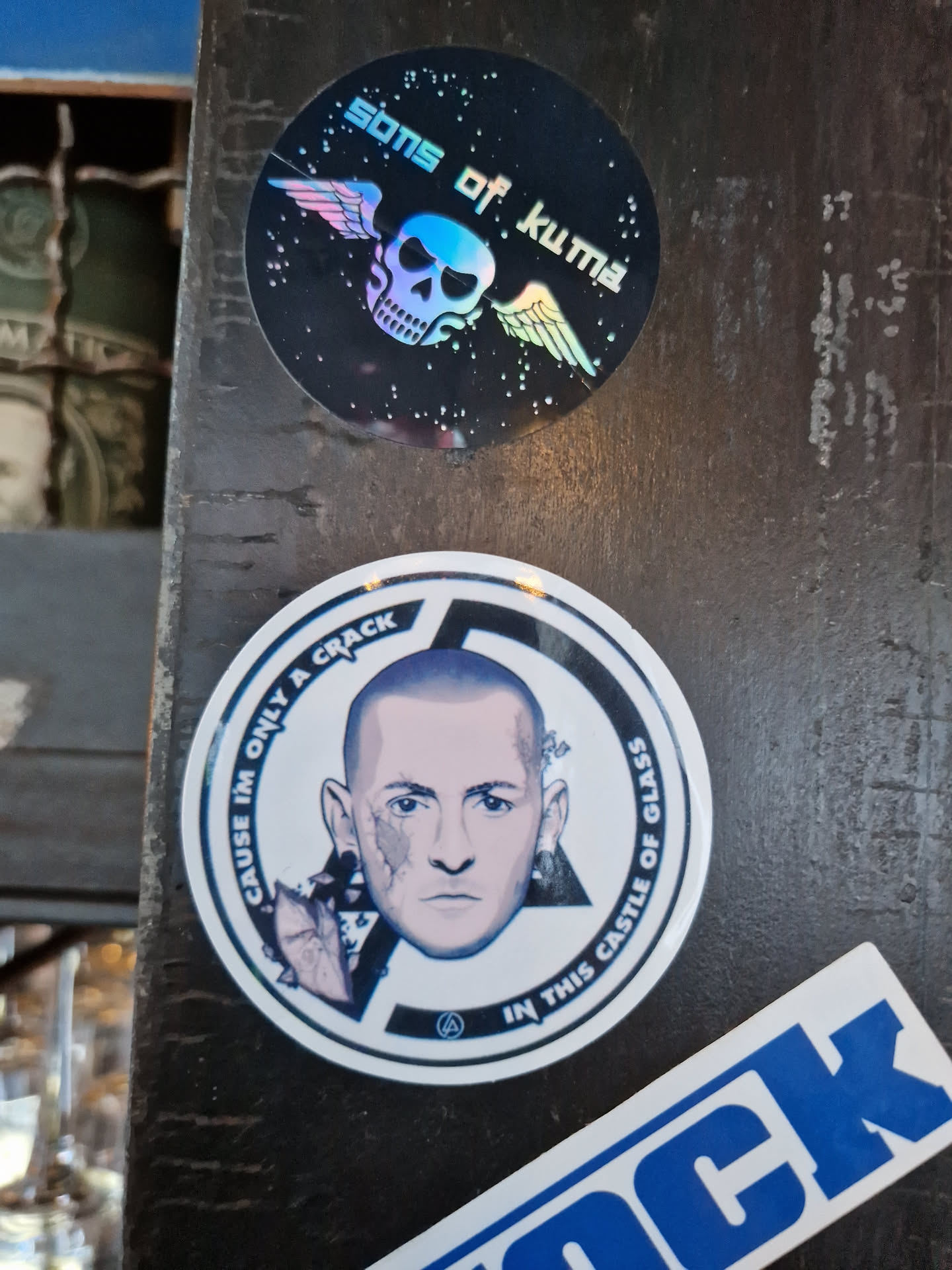 A round sticker featuring a portrait of Chester Bennington, lead singer of Linkin Park. The text surrounding the image reads, "Cause I'm only a crack in this castle of glass."  There is also a smaller sticker featuring a skull with wings and the text "Sons of Kuma".