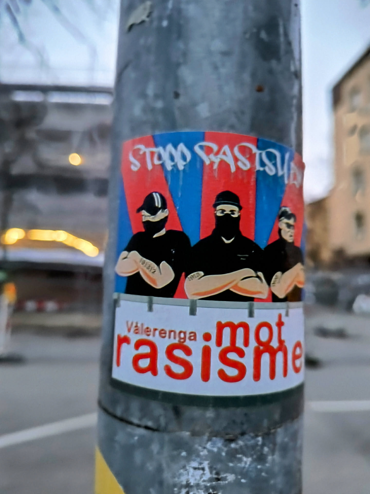 A sticker featuring three figures with arms crossed, wearing black and white, with a background of red and blue stripes and the text "Stopp Rasisme" and "Vålerenga mot rasisme".