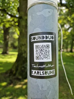 A black and white sticker with the text 'SOUNDDUO', a QR code, 'Technokultur', and 'KARLSRUHE'. The sticker is affixed to a gray metal pole in a park-like setting.