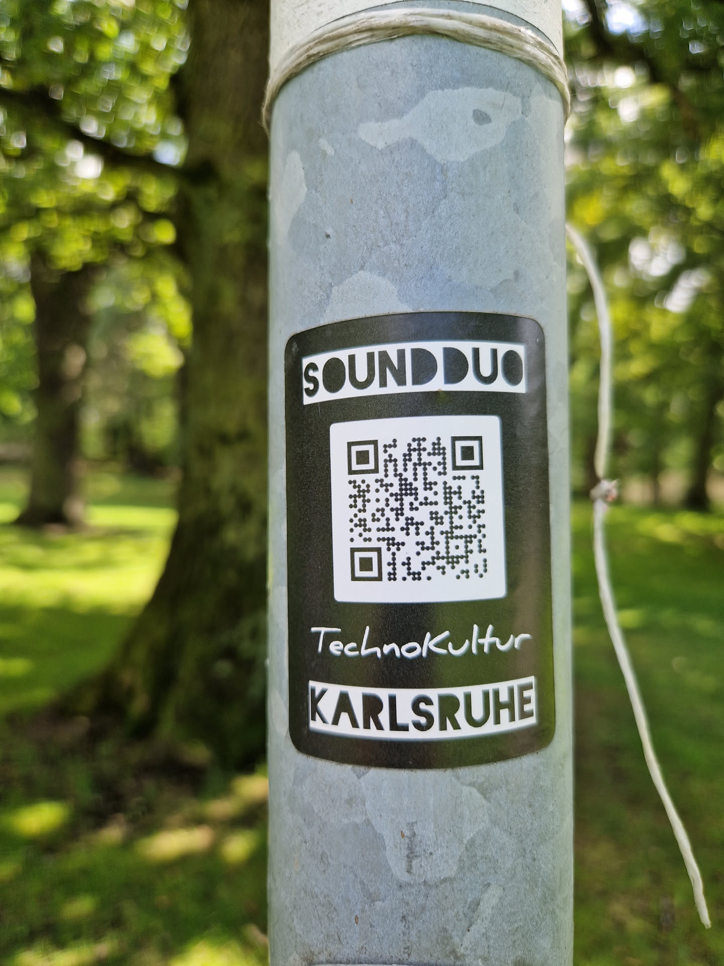 A black and white sticker with the text 'SOUNDDUO', a QR code, 'Technokultur', and 'KARLSRUHE'. The sticker is affixed to a gray metal pole in a park-like setting.