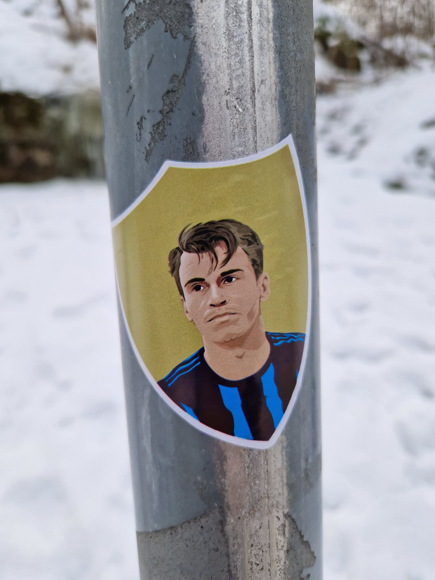 A sticker featuring an illustrated portrait of a soccer player. He's depicted in a retro style, wearing a dark jersey with light blue vertical stripes. The artwork is set within a shield shape on a yellow background. The sticker is affixed to a gray metal pole in a snowy outdoor setting.