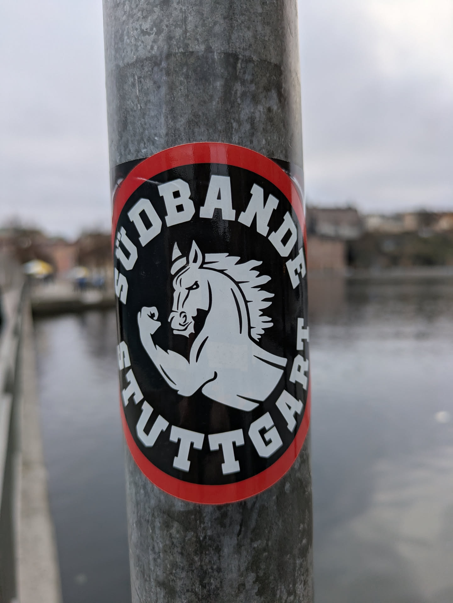 A round sticker with a red border and black background featuring a stylized illustration of a muscular horse.  The text 'SÜDBANDE STUTTGART' is prominently displayed in a circular design around the horse. The sticker appears to be affixed to a metal pole near a body of water.