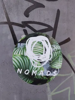 Round sticker with a design featuring tropical leaves and an abstract white scribble. The word "NOMADS" is written at the bottom. It is stuck on a grey wall with graffiti.