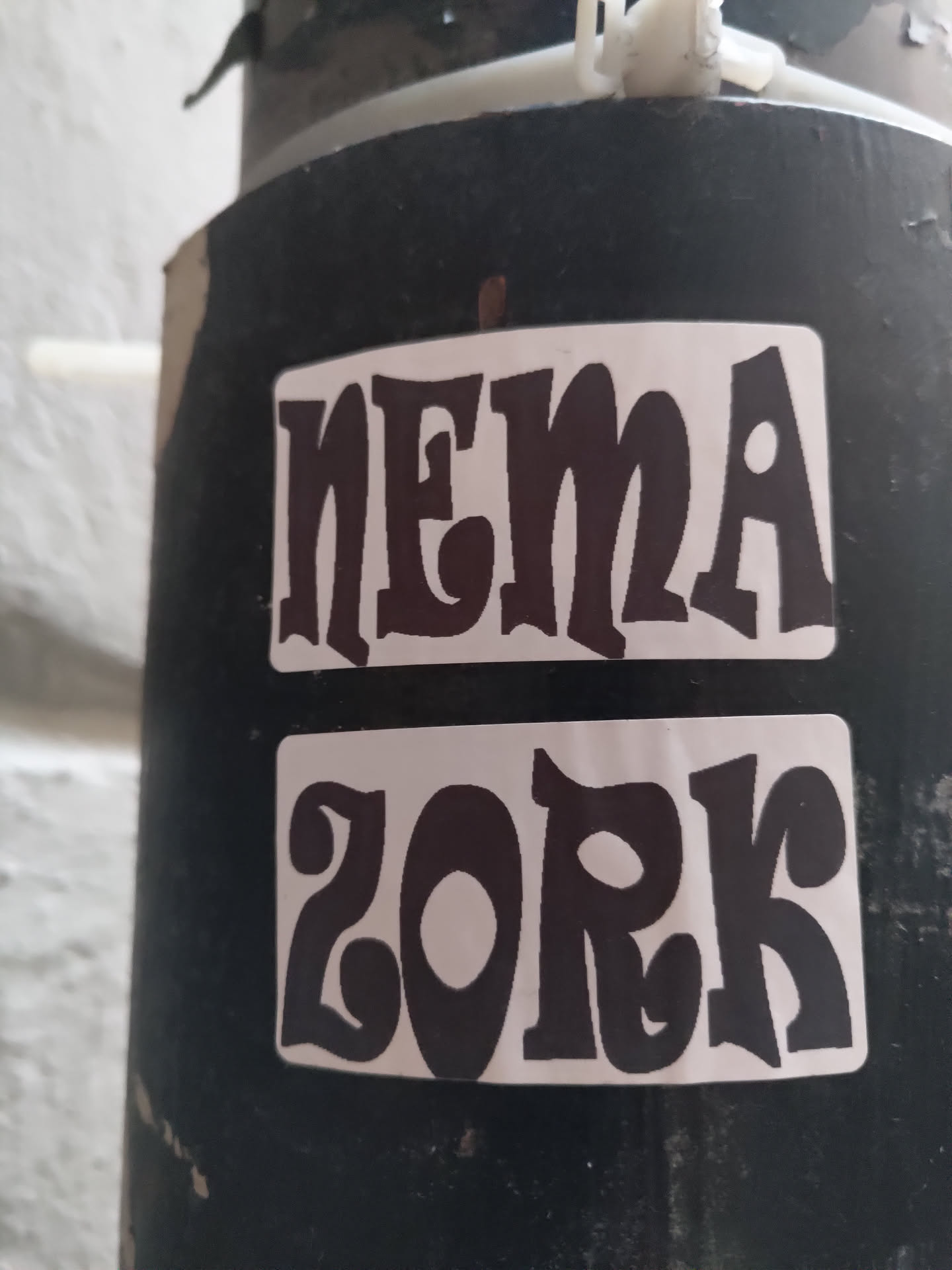 Two-part sticker featuring stylized text. The top sticker reads "NEMA" and the bottom sticker reads "ZORK". Both are in black text on a white background with a retro font style.  The stickers are affixed to a dark-colored pole outdoors.