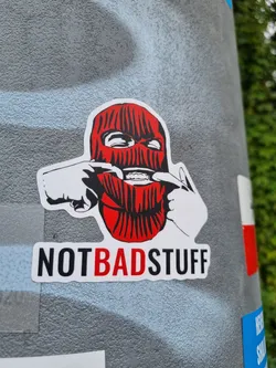 A sticker featuring a stylized illustration of a person wearing a balaclava, pulling their own cheeks. The text "NOTBADSTUFF" is written below the image. The style is bold and graphic, with a slightly rebellious tone.