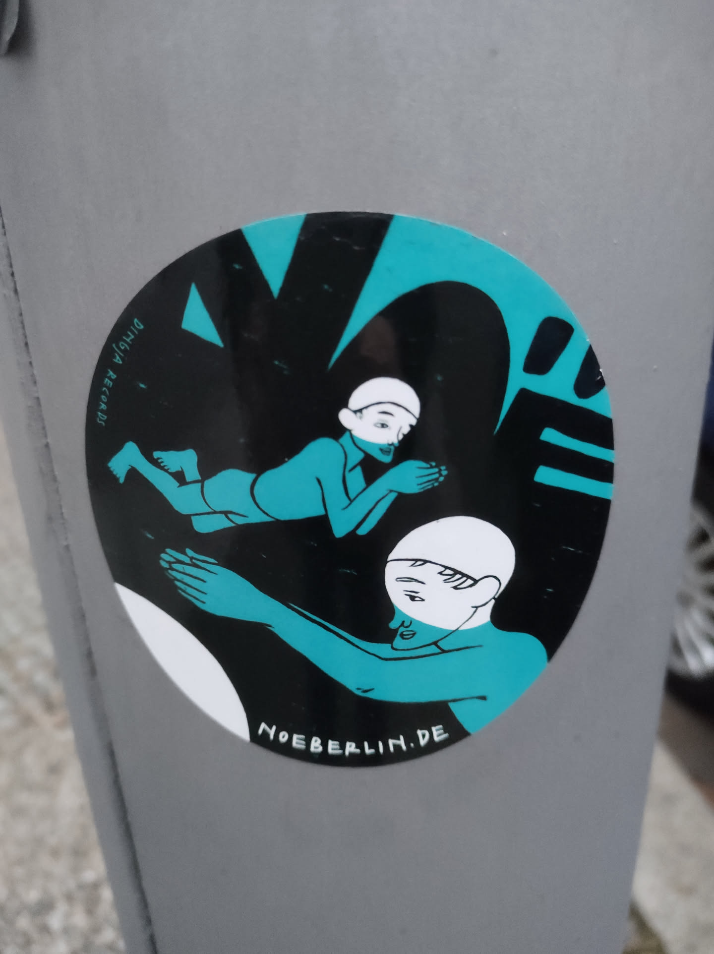 Round sticker featuring a minimalist, stylized illustration in teal and black. Two figures are depicted, one seemingly reclining and the other reaching towards them. The artwork has a bold graphic style. The text 'NOEBERLIN.DE' is visible at the bottom.