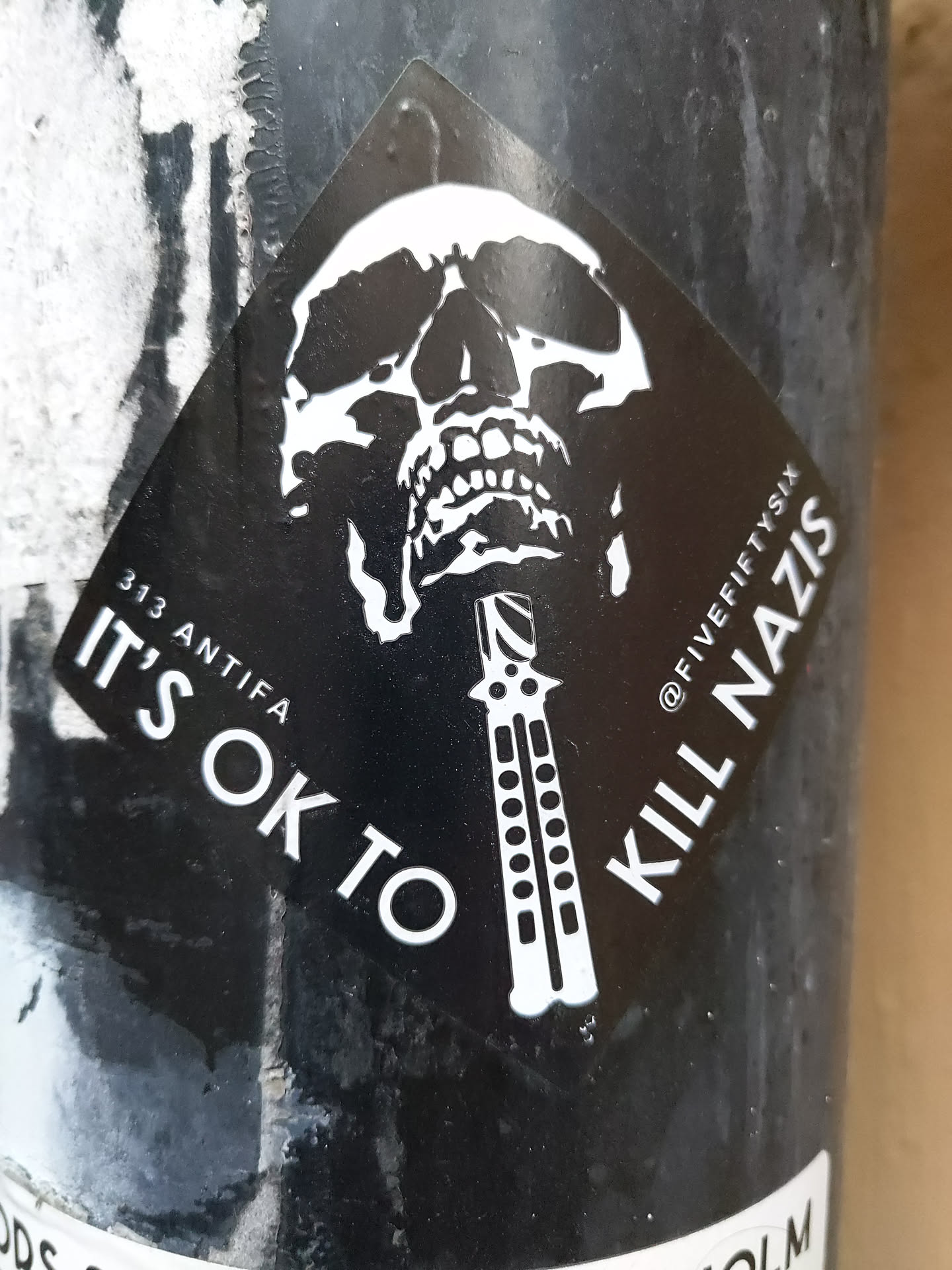 A black and white sticker with a skull and a switchblade. The text on the sticker says "It's ok to kill nazis". There is also a small text on the top right that says @fivefiftysix and 313 antifa on the bottom left. 
