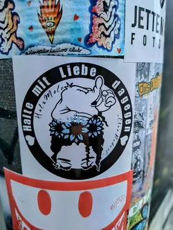 A black and white sticker with a illustration of a woman with sunflowers in her hair giving a thumbs up. The text says "Halte mit Liebe dagegen" which translates to "Stand up against with love".
