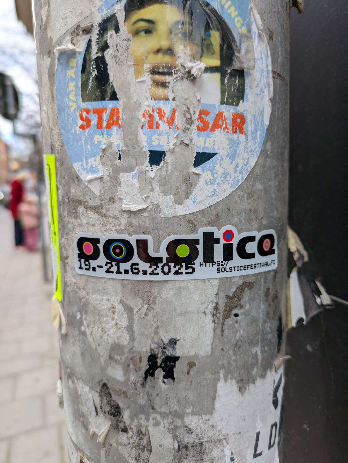 This is a sticker advertising the Solstice Festival which will occur from June 19th to June 21st, 2025. The sticker is on a white background with a black border and features colorful circles within the word Solstice. It is attached to a light grey pole with various other stickers and posters.