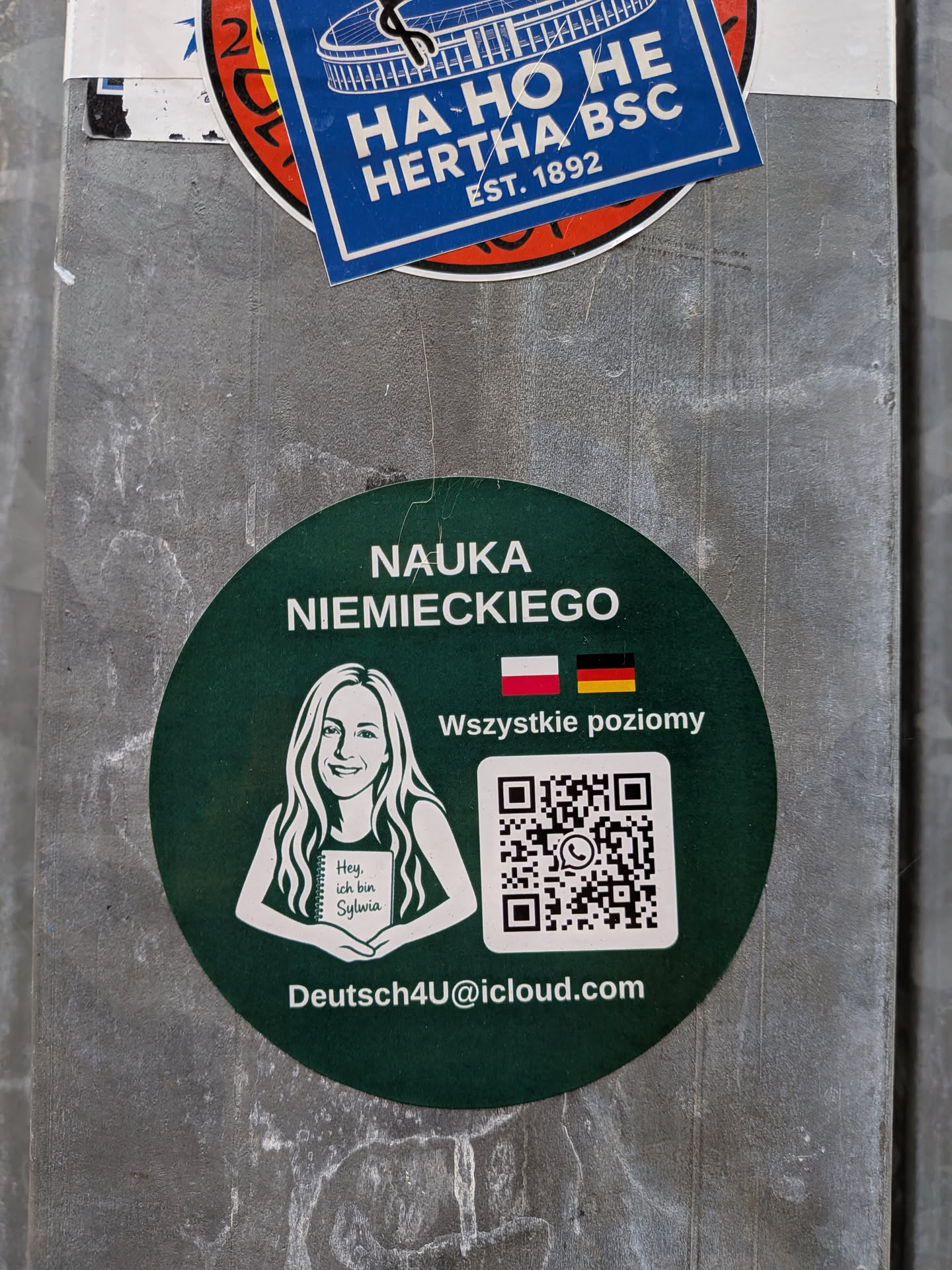 A circular dark green sticker advertising German language learning. At the top, bold white text reads 'NAUKA NIEMIECKIEGO' (Learn German). On the left is a black-and-white line illustration of a smiling woman holding a notebook that reads 'Hey, ich bin Sylwia'. To the right are the national flags of Poland and Germany followed by the Polish phrase 'Wszystkie poziomy' (All levels), and a white square containing a QR code centered with a WhatsApp icon. Along the bottom edge, the contact email 'Deutsch4U@icloud.com' is printed in white.