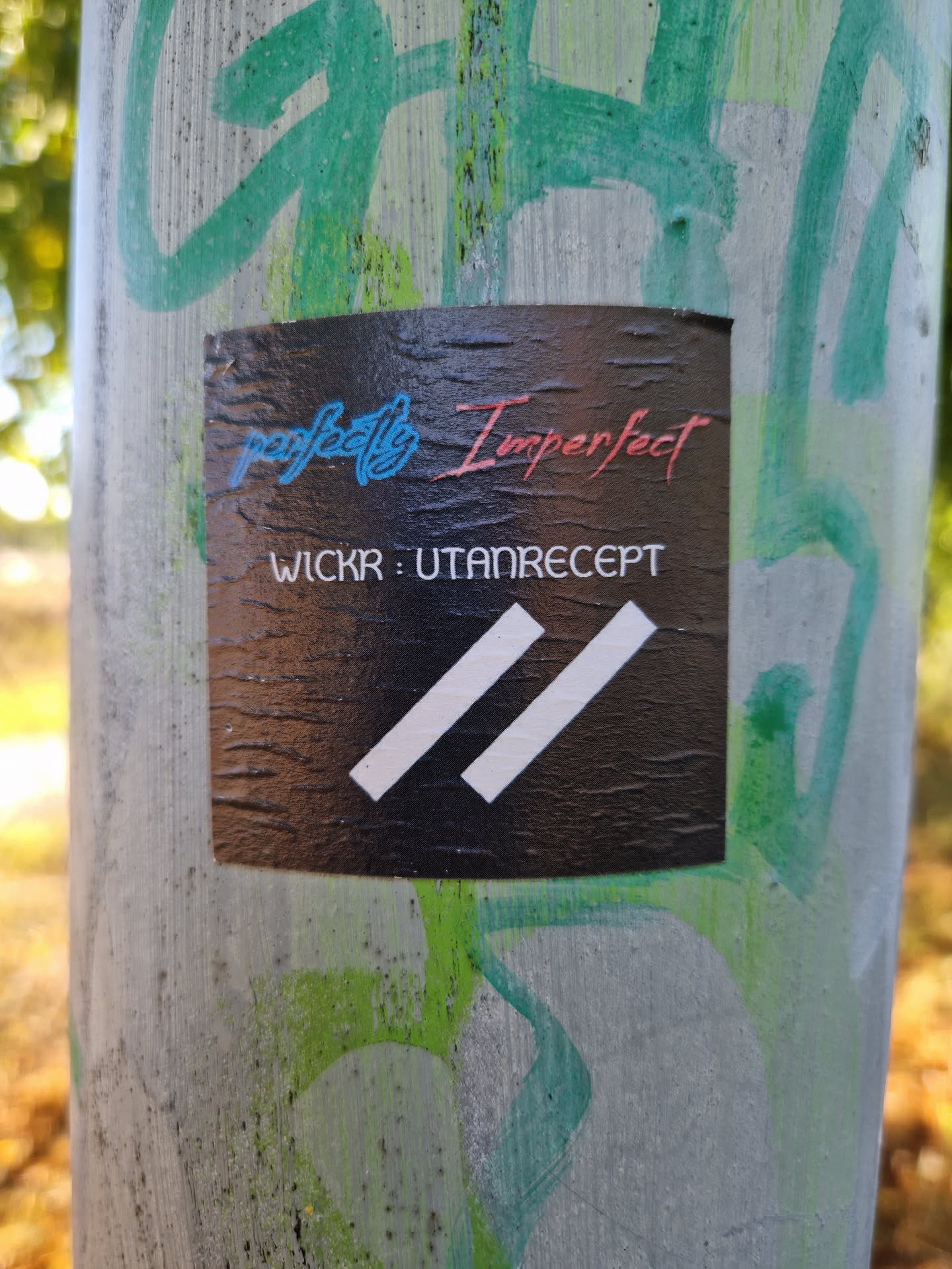 Black sticker with the text "perfectly Imperfect" in a stylized font and the Wickr contact information: "WICKR: UTANRECEPT".  It also features two parallel white stripes at the bottom. The sticker appears to be affixed to a pole with some graffiti in the background.