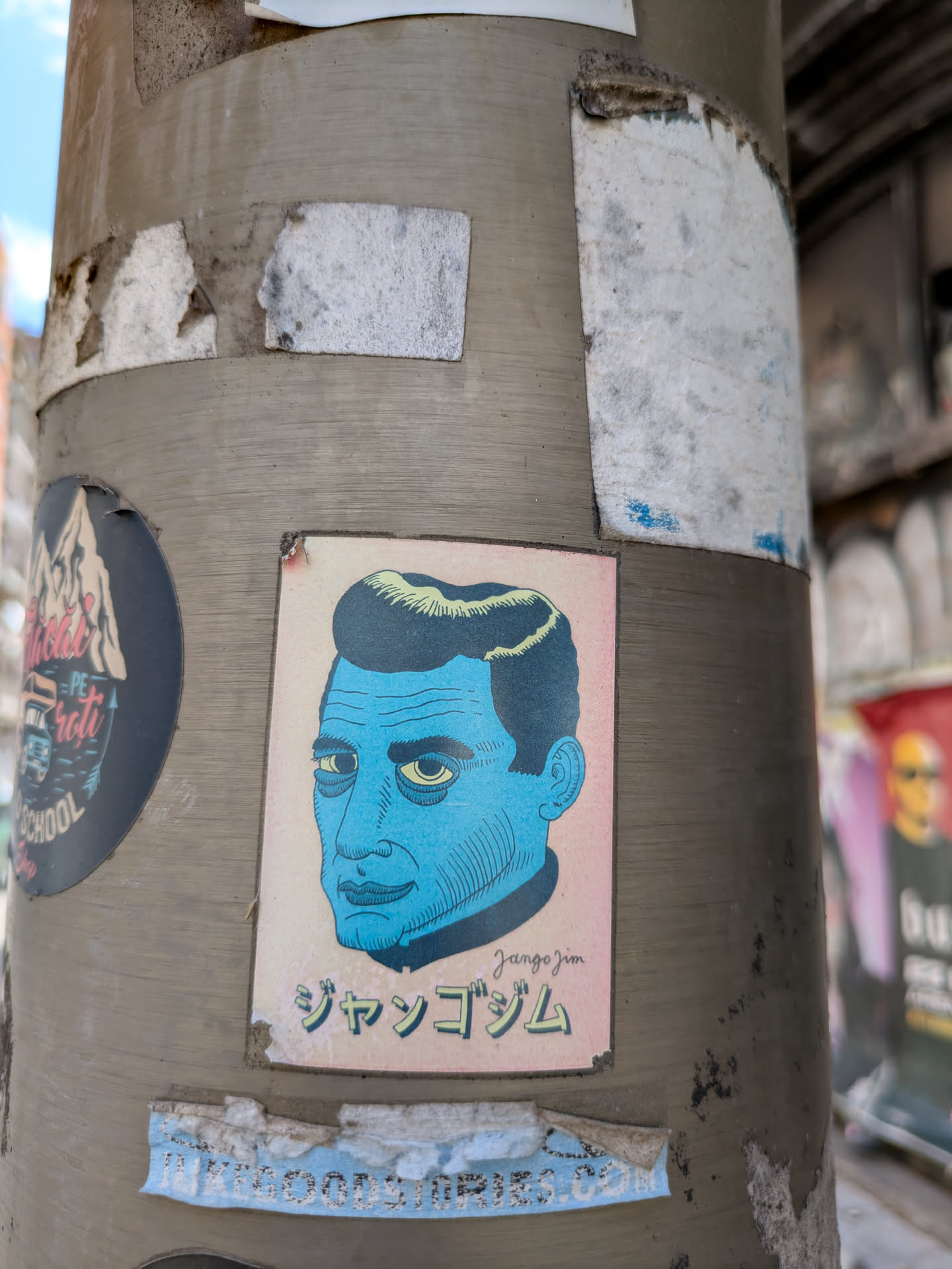 A blue-toned sticker of a man's face is affixed to a gray pole. The sticker, which is slightly worn, features a man with sharp features and yellow eyes. Below the portrait, the text "jangojim" is written in a stylized font, possibly in Japanese.