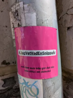 A pink sticker is adhered to a metal pole. The sticker contains white text that reads "#JagVetVadEnSnippaÄr och den som inte gör det ska inte sitta i en domstol", promoting awareness.