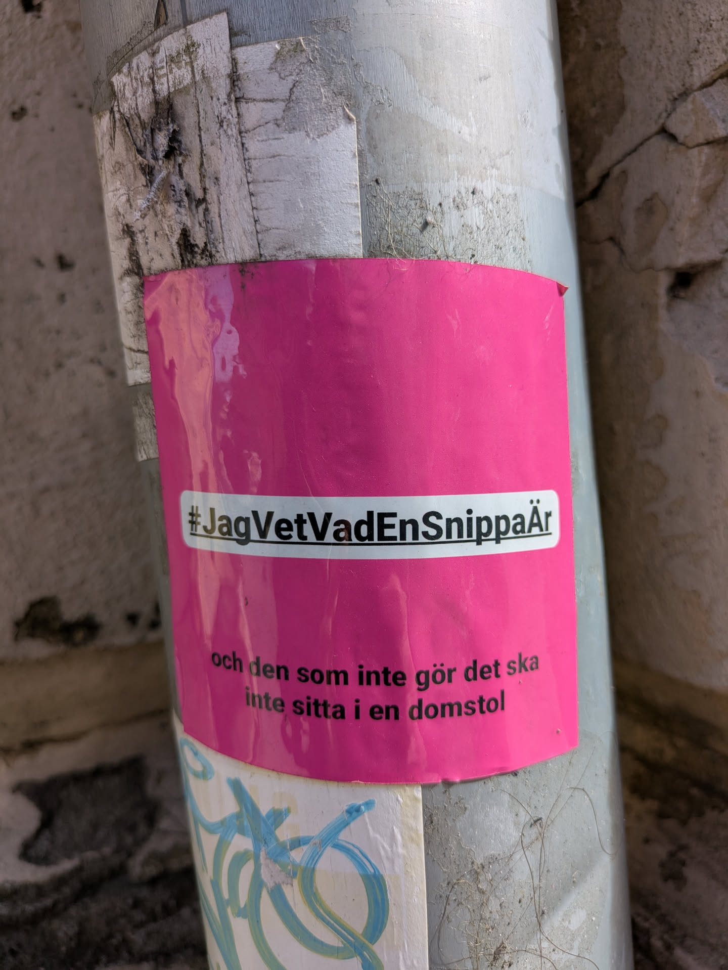 A pink sticker is adhered to a metal pole. The sticker contains white text that reads "#JagVetVadEnSnippaÄr och den som inte gör det ska inte sitta i en domstol", promoting awareness.