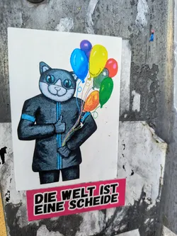 This sticker depicts an illustration of a cat dressed in a suit and holding a bunch of colorful balloons. Beneath the cat is a text in German language. The overall impression is a mix of whimsy and bold statement.