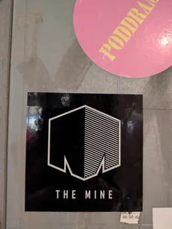 This is a black square sticker with a white logo on it. The logo consists of an abstract shape resembling a partially mined structure. The text "THE MINE" is printed below the logo.