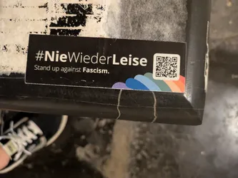 The sticker is black with white text and a small rainbow graphic. A QR code is located on the right side of the sticker.
